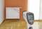 An infrared thermometer with laser beams measures the temperature of a radiator in a room with a wooden floor and orange wall.