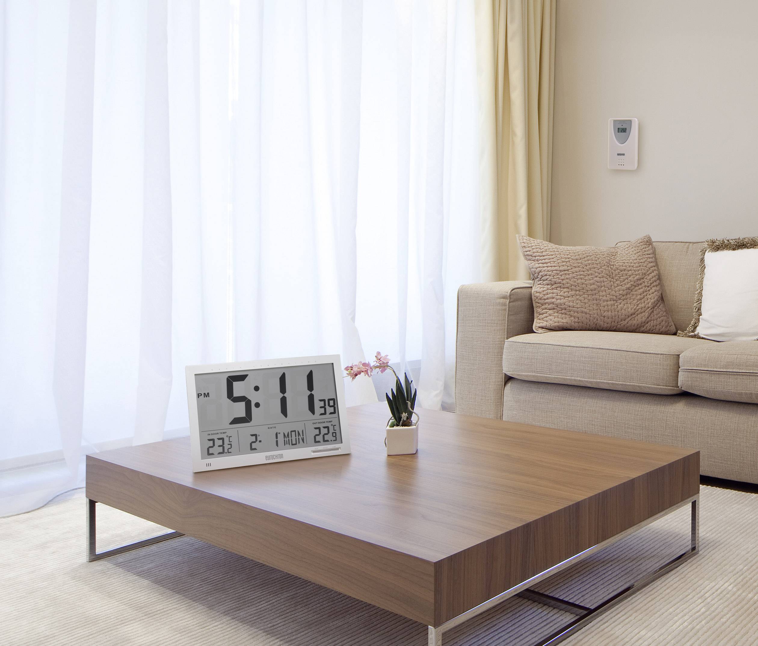 A modern living room with a wooden table, on which a large digital alarm clock sits. A sofa with cushions is in the background.
