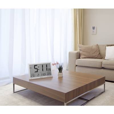 A modern living room with a wooden table, on which a large digital alarm clock sits. A sofa with cushions is in the background.