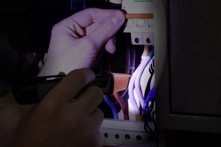 A hand holds a torch, illuminating an open electrical panel. Several colourful cables are visible.