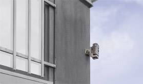 A grey building facade with windows on the left side, a small camera mounted on the wall and pointing to the right.