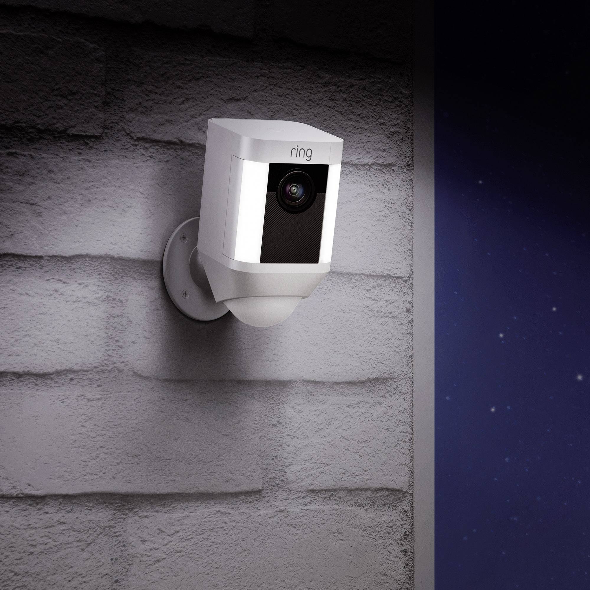 A white security camera on a grey wall in an outdoor area, possibly at night, as the background is dark.