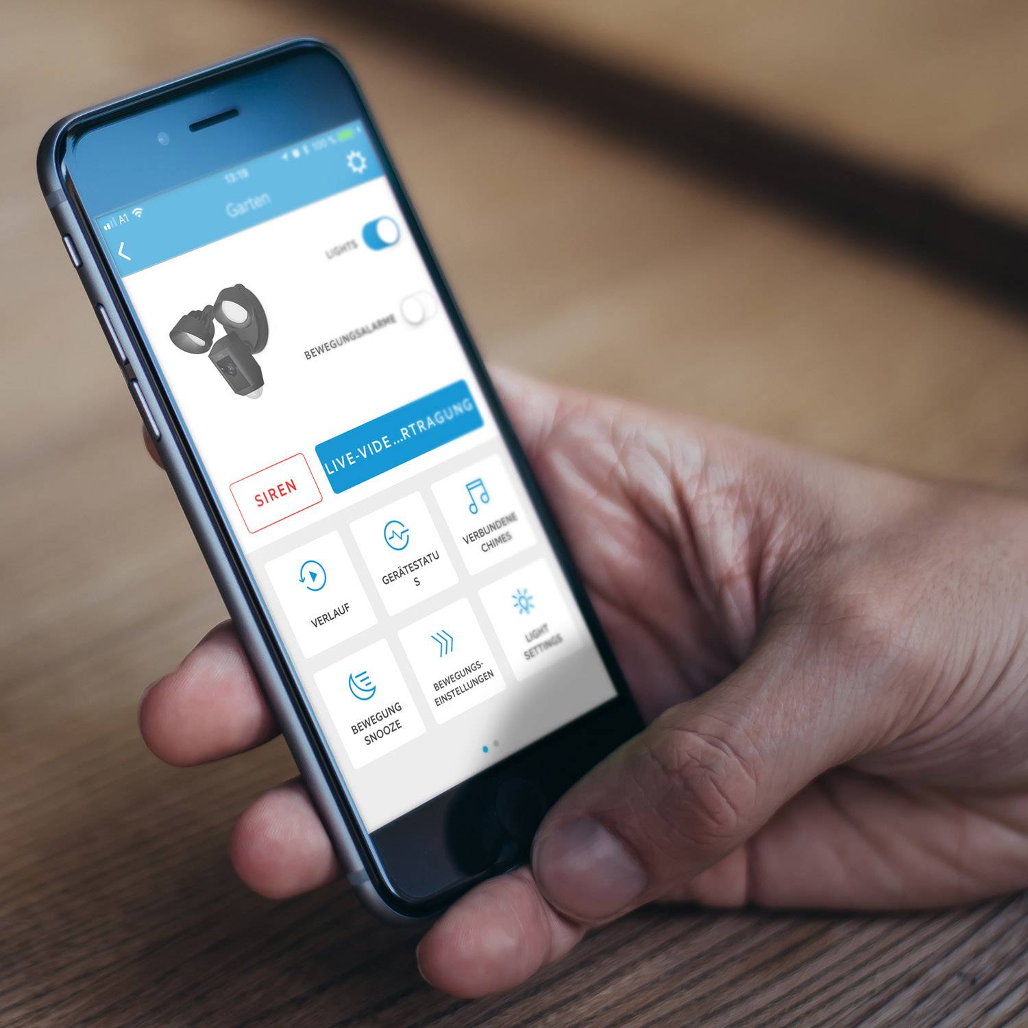 A hand is holding a smartphone with an open security app. The app displays options such as 'Siren', 'Remote Control', and 'Settings'.