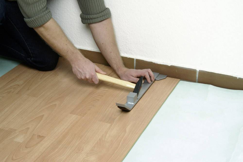 A person is laying laminate flooring and using a pull bar and hammer to secure the boards tightly together.