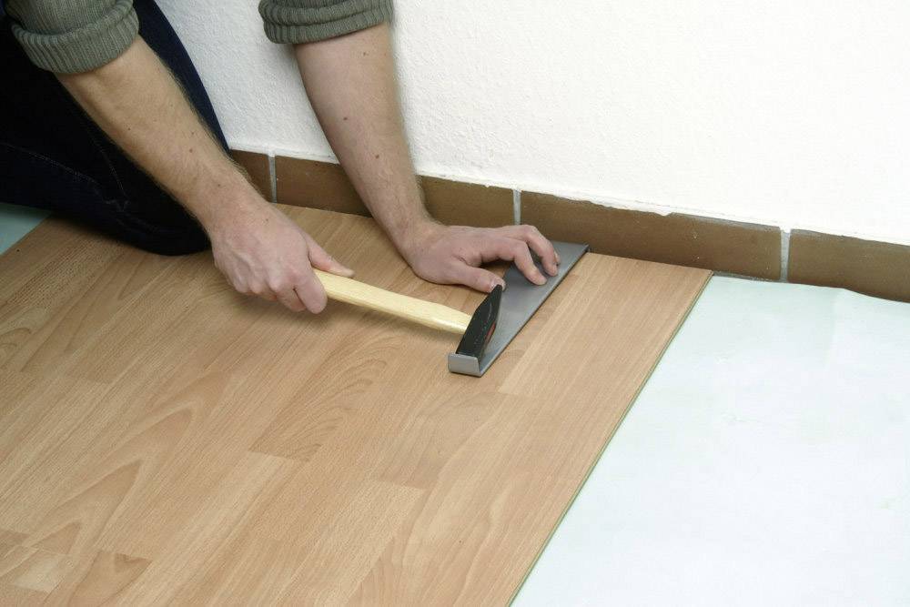 A person is laying laminate flooring and using a tapping block and hammer to join the panels tightly together.