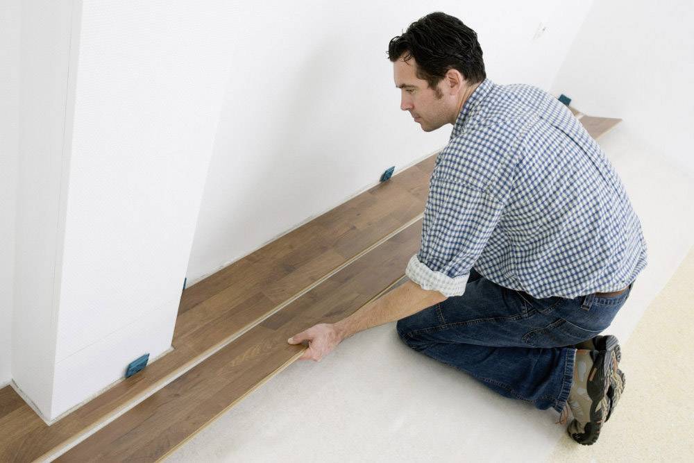 A man is laying laminate flooring in a room. He is kneeling on an underlay and fitting the wooden boards against the wall.