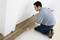 A man is laying laminate flooring in a room. He is kneeling on an underlay and fitting the wooden boards against the wall.