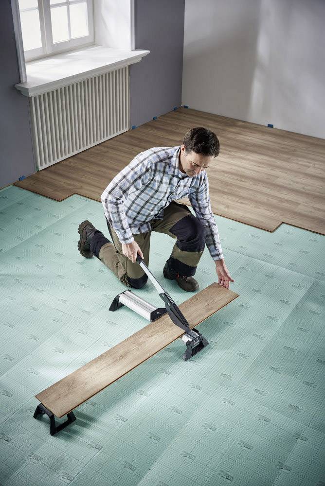 A man is laying laminate flooring. He is using a tool to cut the planks. The room has a green underlay and wood-effect décor.