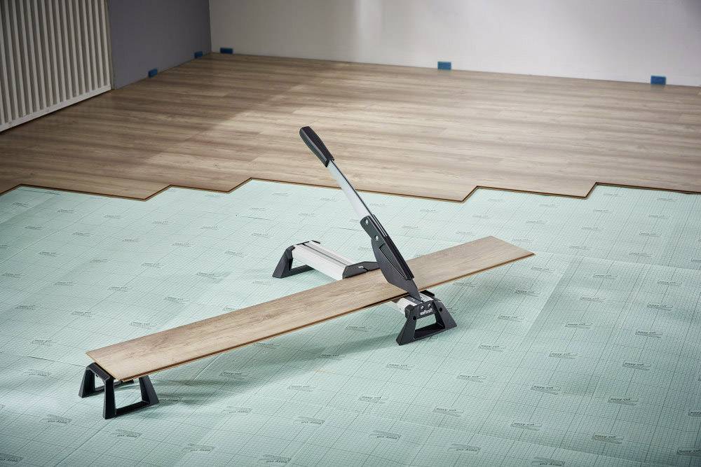 A laminate cutter is lying on a partially laid laminate floor. The room shows an ongoing floor installation.