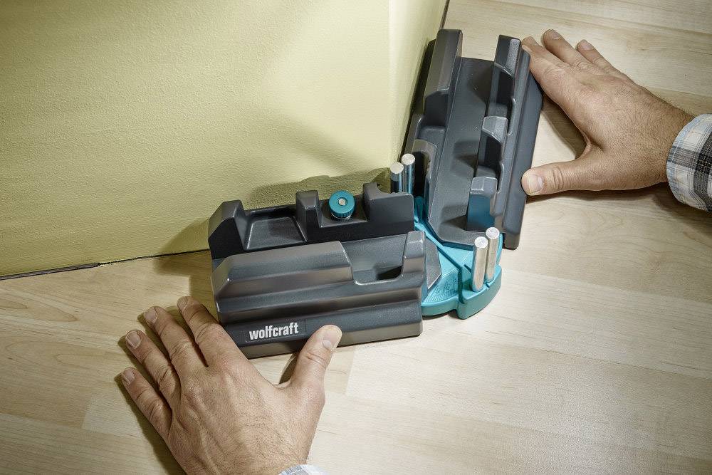 A tool for measuring angles and corners on a wall. One person is aligning the device at a corner on a wooden floor.