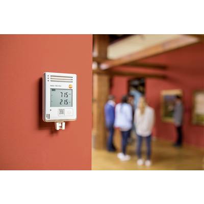 A digital temperature and humidity metre mounted on a red wall displays '21.5°' and '71.5%'. People can be seen in a room in the background.