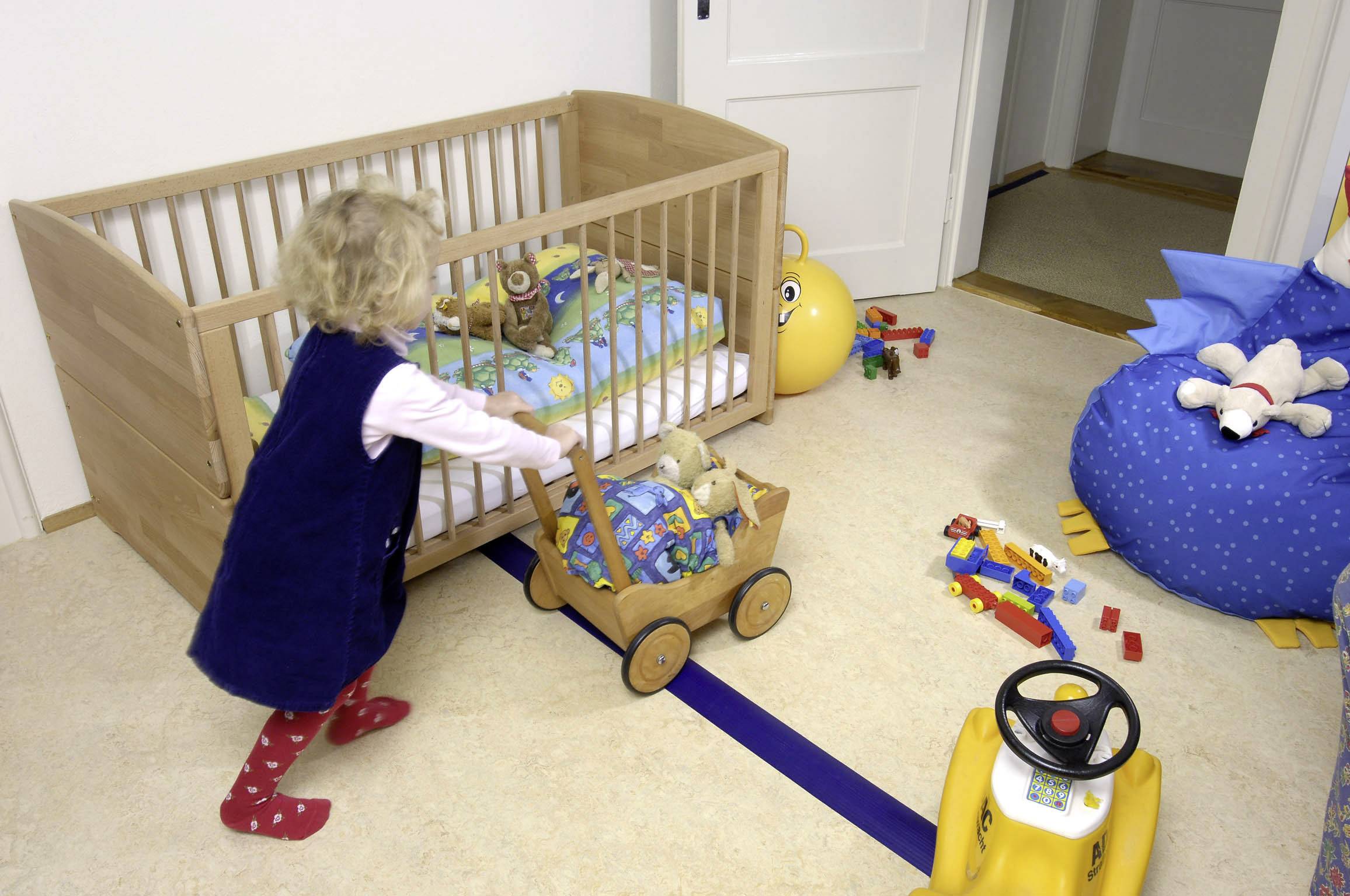A small child is playing in a children's bedroom. They are pushing a wooden toy cart. In the background, a cot stands.