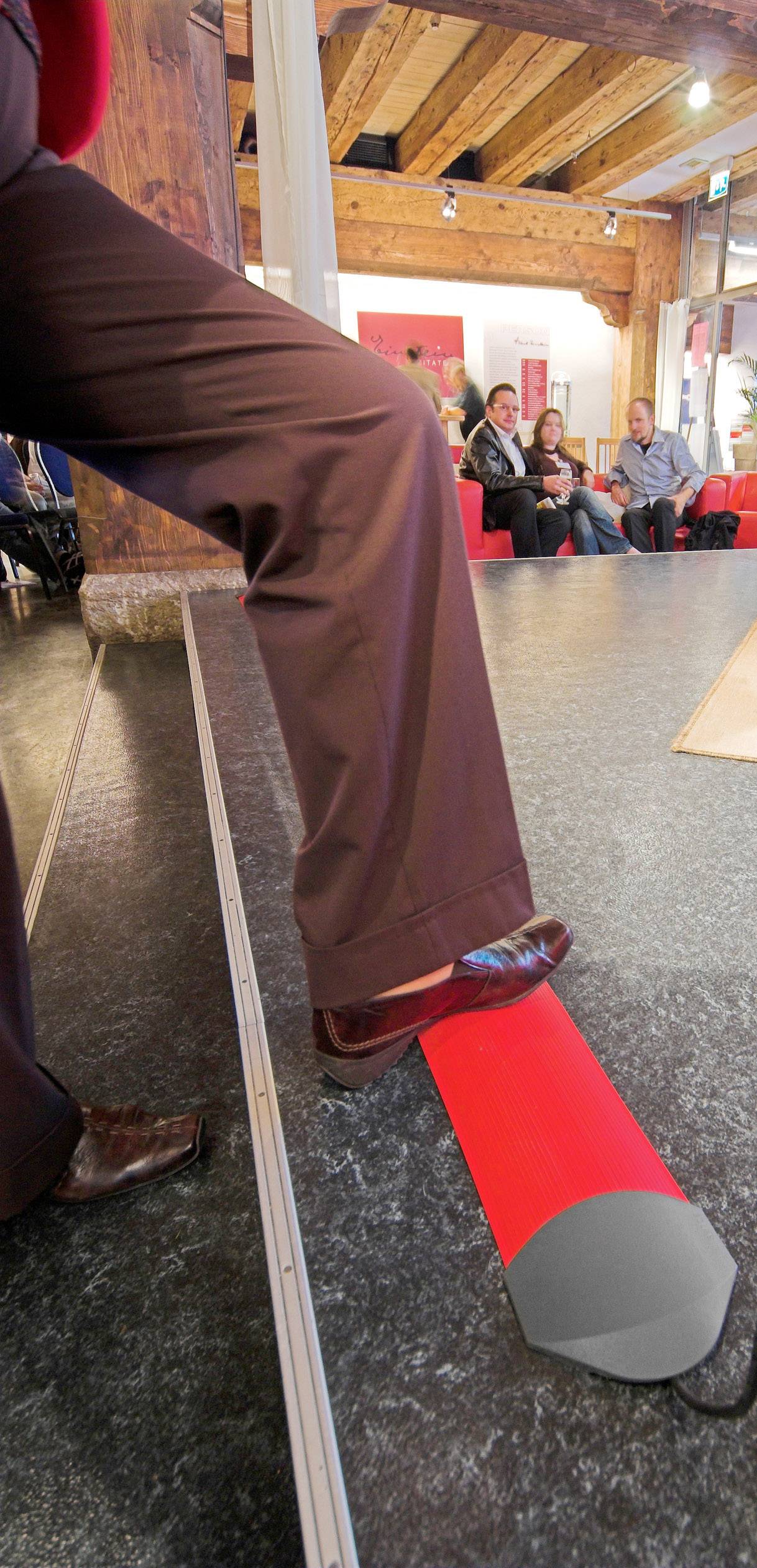 A person wearing brown trousers is standing on a red cylinder in a room with a wooden beam ceiling. In the background, people are sitting on red chairs.