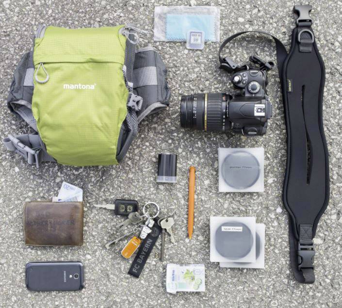 Camera equipment spread out on asphalt, consisting of a rucksack, DSLR camera, lens caps, wallet, bunch of keys and smartphone.