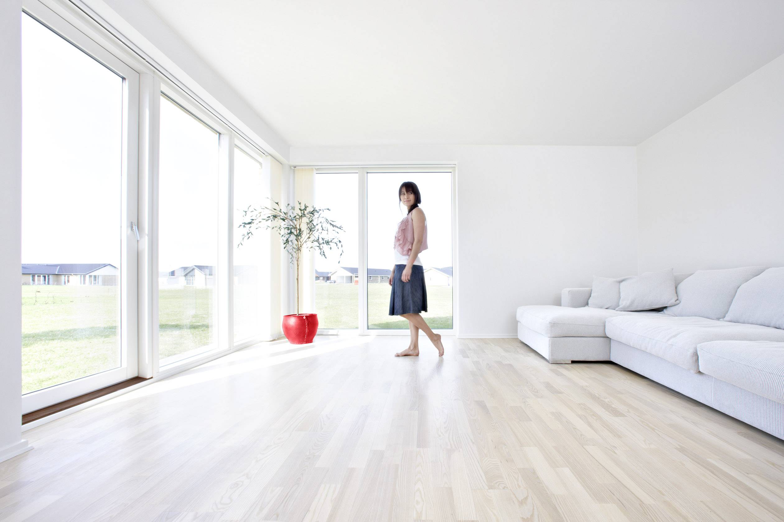 A woman is standing in a large, bright living room with wooden flooring. Large windows allow plenty of light to enter. A plant is positioned on the left.