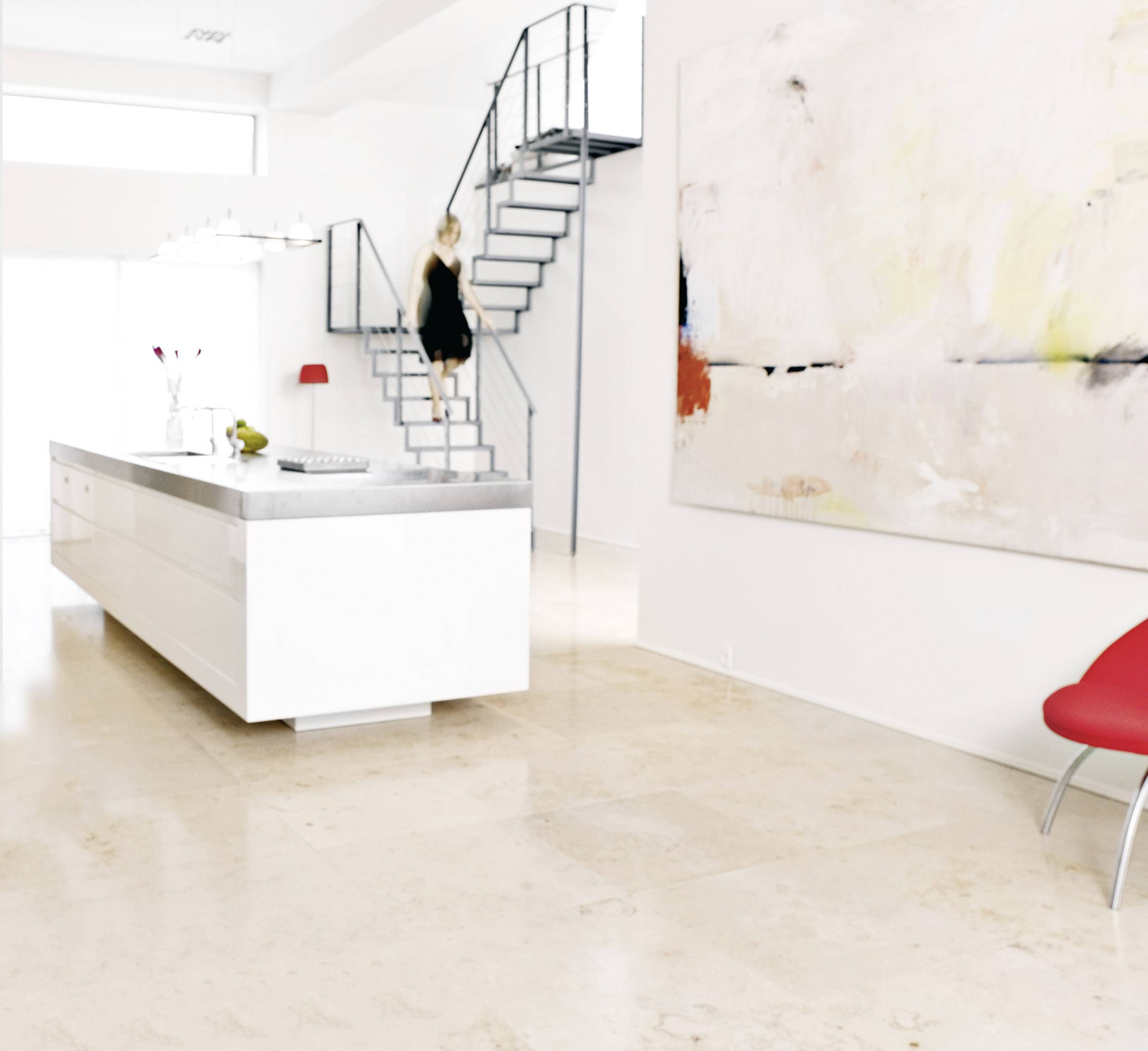 A modern, bright kitchen interior with a central kitchen island, a woman on a staircase to the left and an abstract painting on the wall.