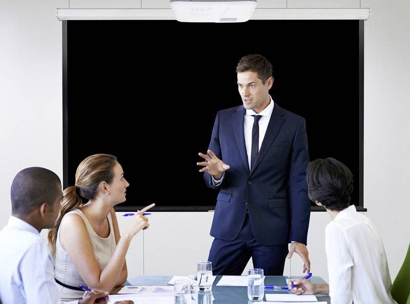 A businessman in a suit is presenting in front of a projection screen, while three listeners at the table watch and take notes attentively.