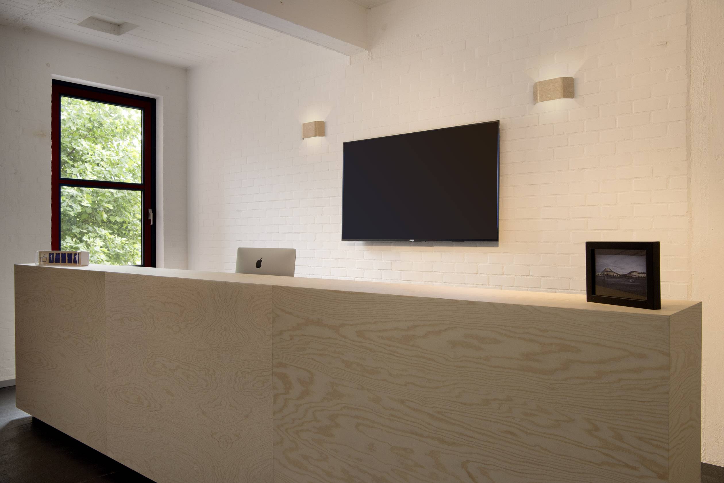A modern reception area with a large, bright wooden desk. A flat-screen monitor and a picture are hung on the white wall.