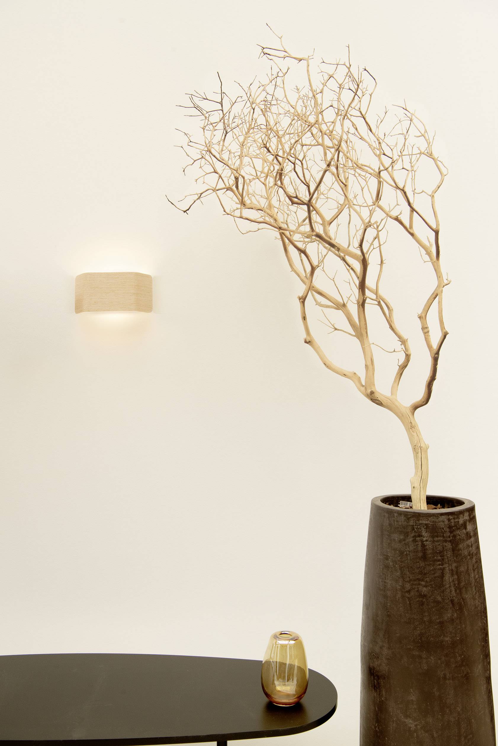 A bare branch in a tall vase stands on a black table. Beside it, a wall lamp is switched on, emitting warm light.