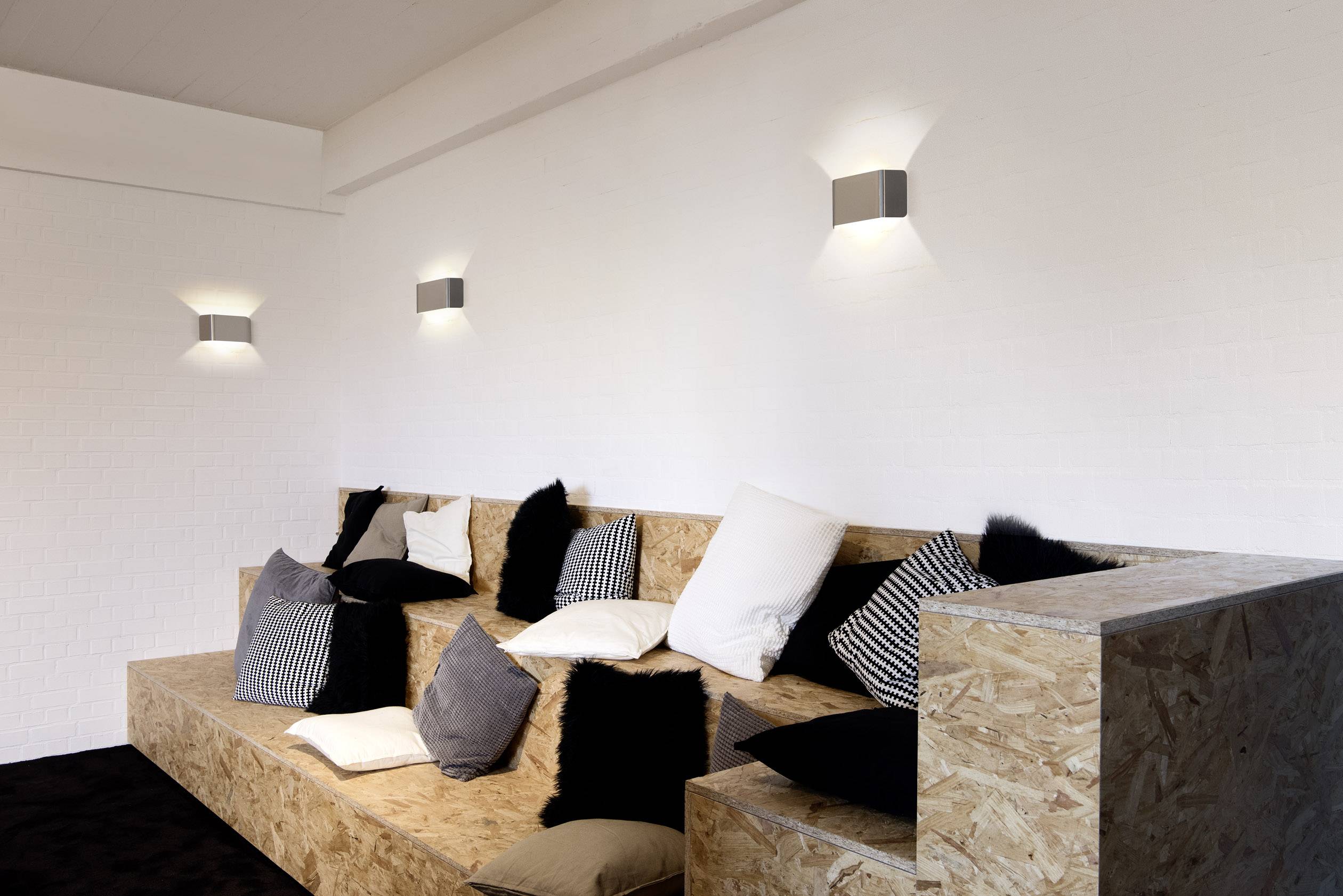 Multilevel wooden seating platform with various cushions on one side of a bright room, wall lights above.