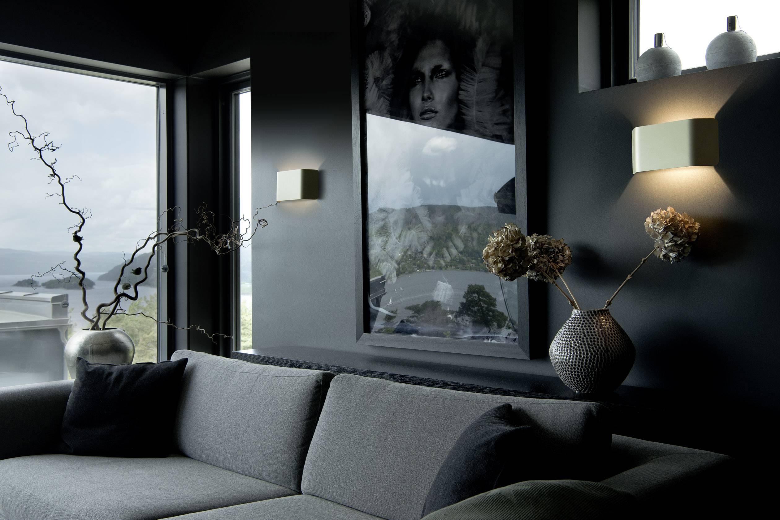 A modern living room with dark walls and a large window offering a view of a landscape. A large, grey sofa sits in the foreground.