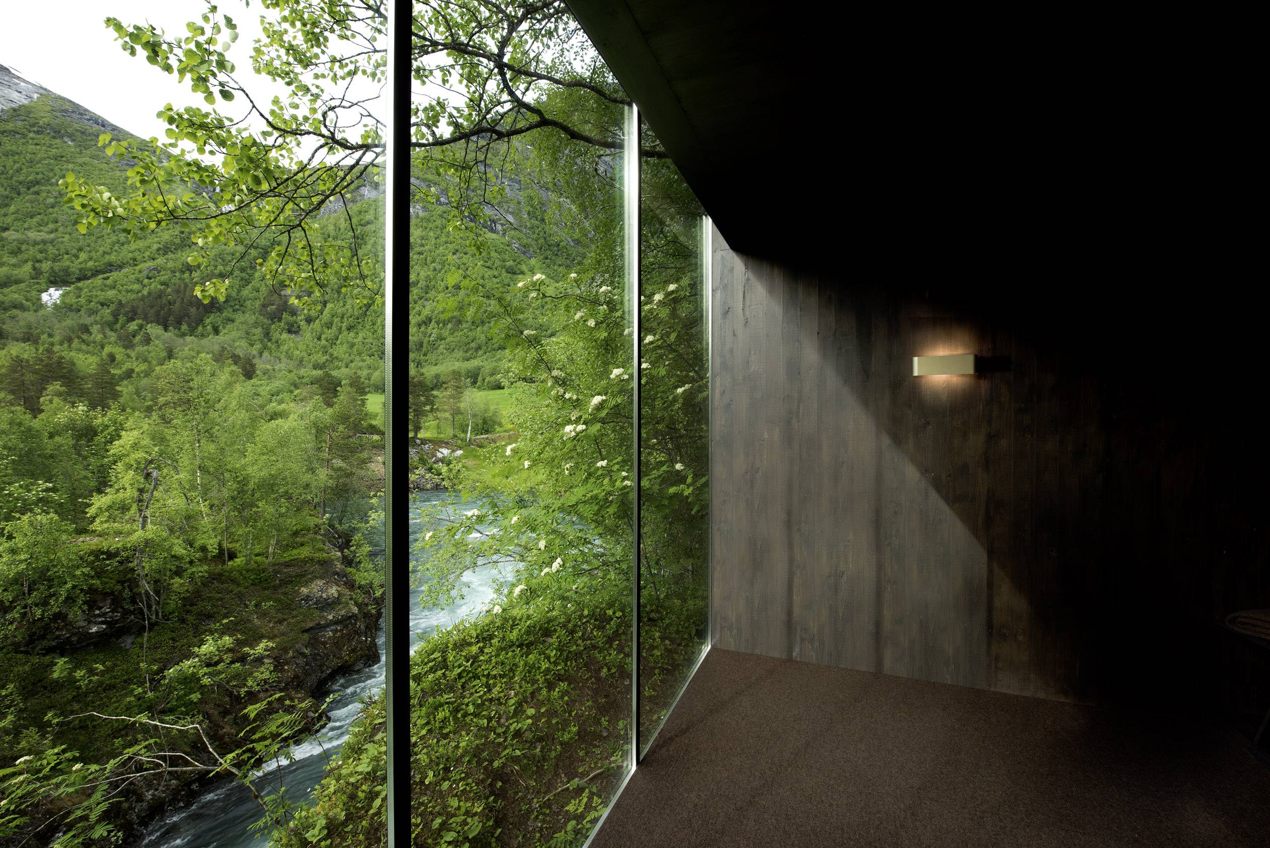 A modern room with large glass walls reveals a view of a lush forest and a river, connecting nature with architecture.