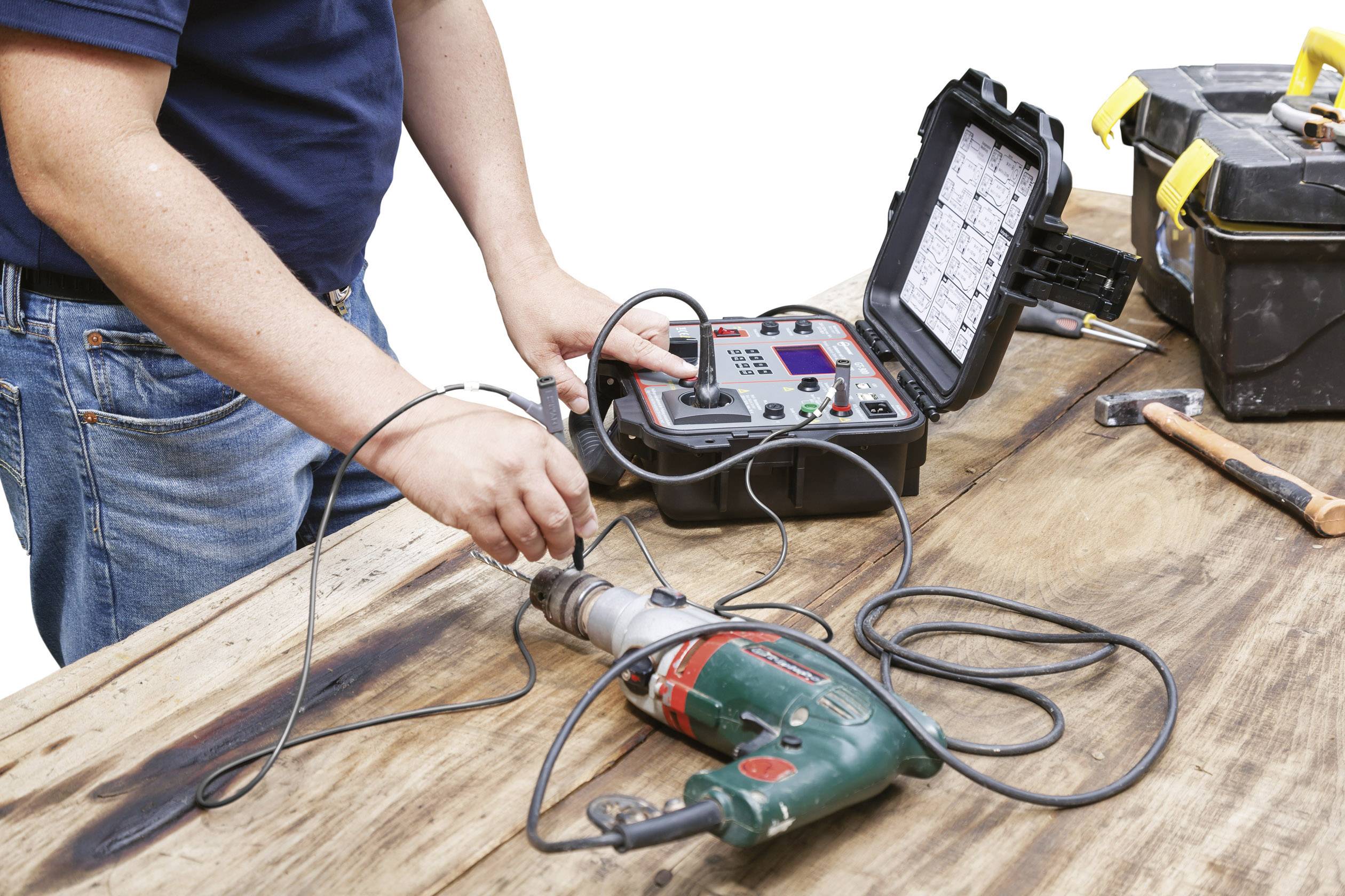 A person is testing an electrical device with a portable measuring instrument on a wooden table. A toolbox is lying nearby.