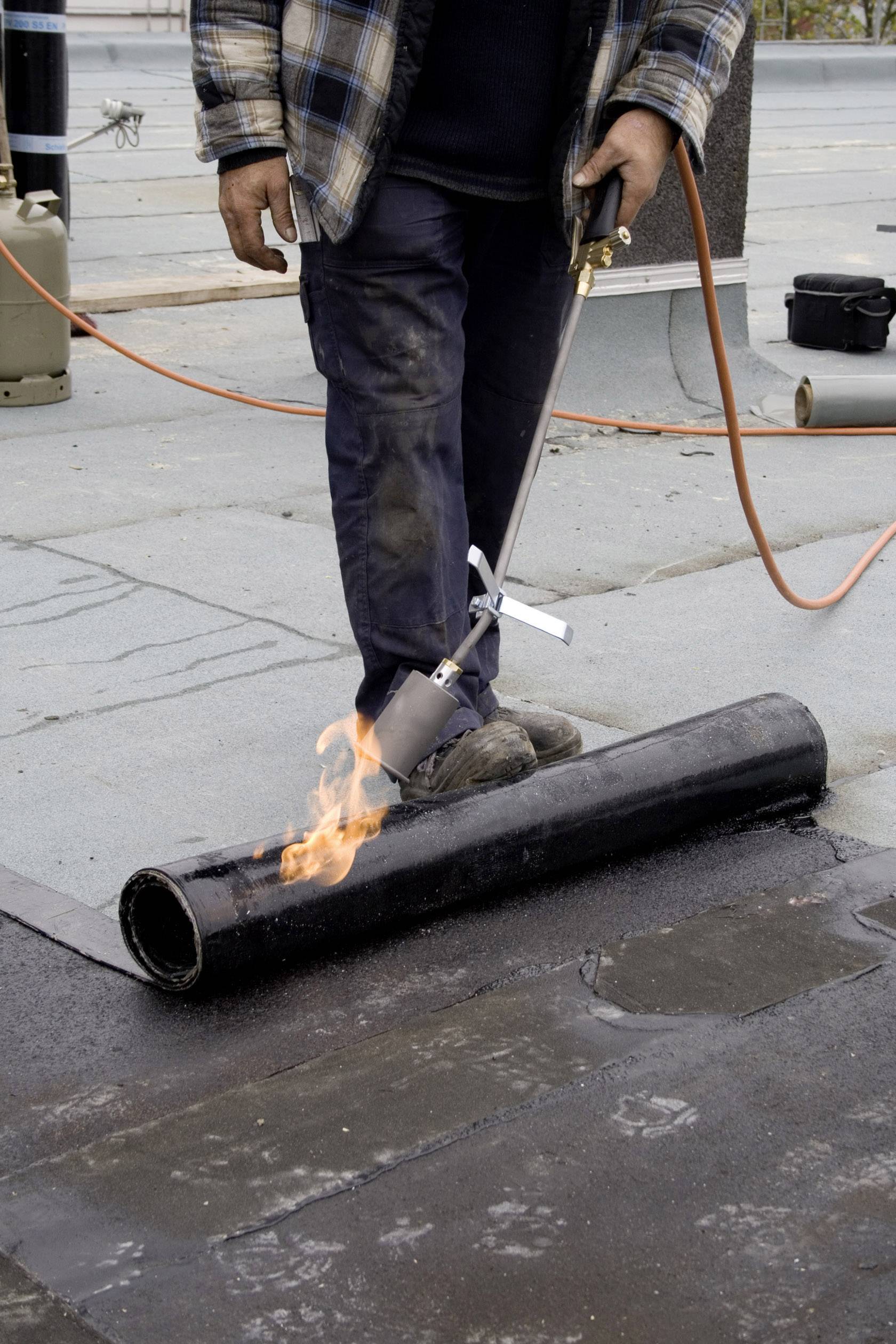 A worker is applying a flame with a burner to roofing felt to bond it together. It is a construction site scene on a flat roof.