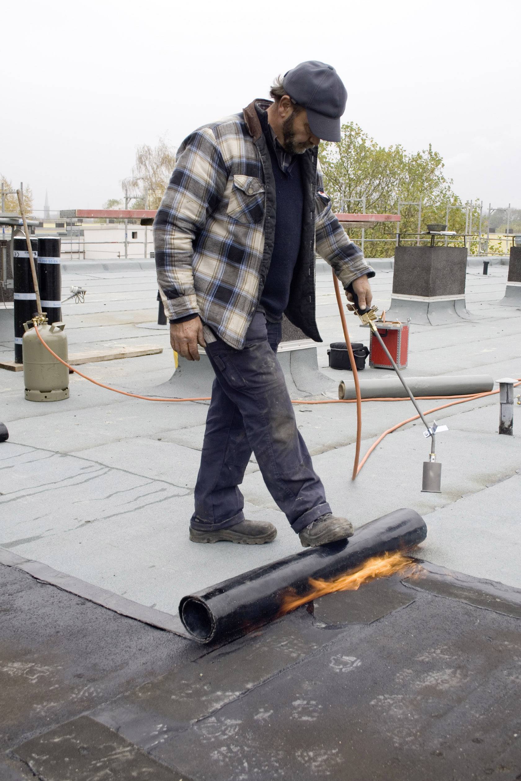 Worker using a blowtorch to secure roofing felt on a flat roof. Weather is cloudy, work area is open.