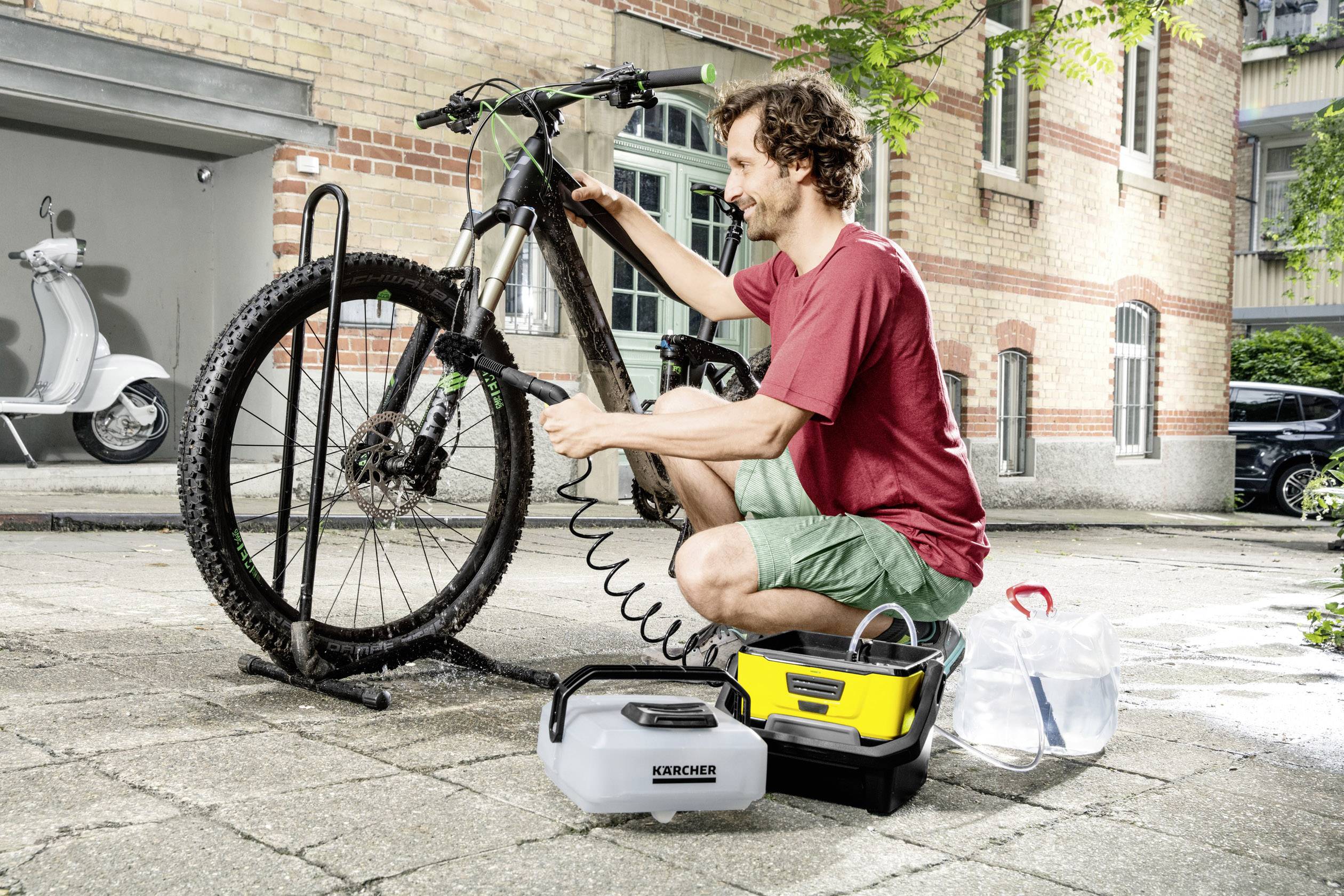 A man is cleaning a mountain bike with a portable pressure washer on a paved courtyard in front of a brick building.