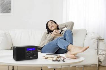 A woman is relaxing on a sofa with a mug in her hand. Next to her, a switched-on radio sits on a table.