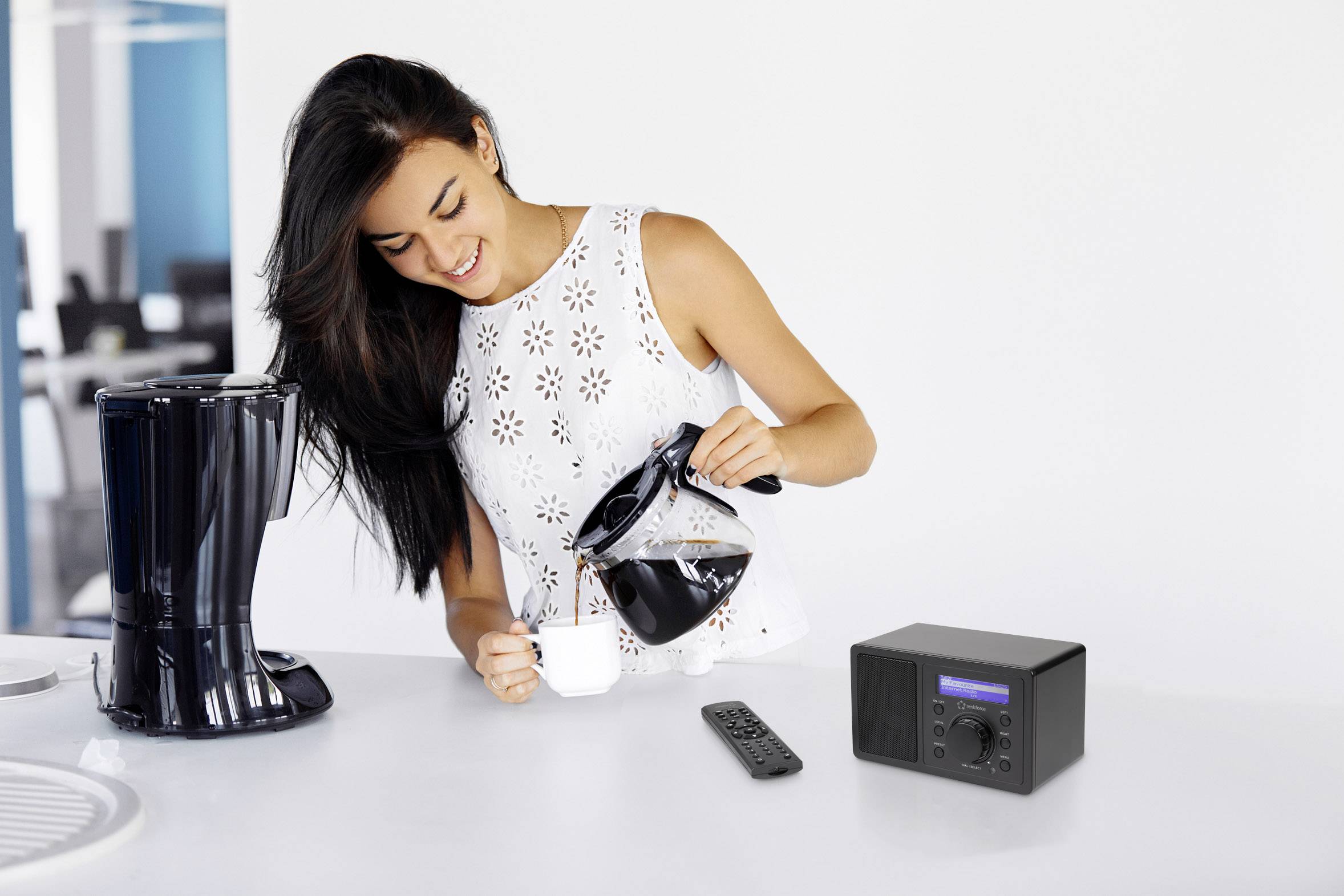 A woman pours coffee into a cup next to a coffee machine; a remote control and a small radio are lying on the table.