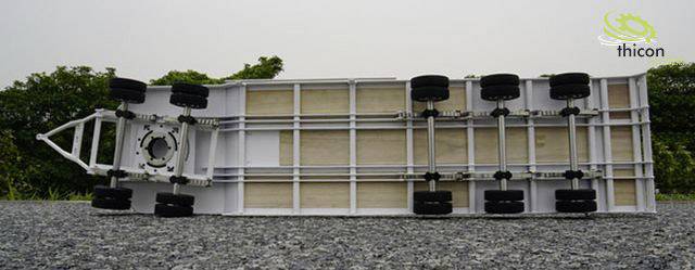 Underside of a model truck chassis with multiple wheels, photographed on a gravel path, surrounded by trees in the background. Logo in the top right corner.