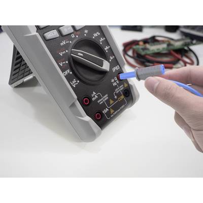 A person is using a digital multimeter on a table. The scale shows various measuring ranges. A blue cable is connected.