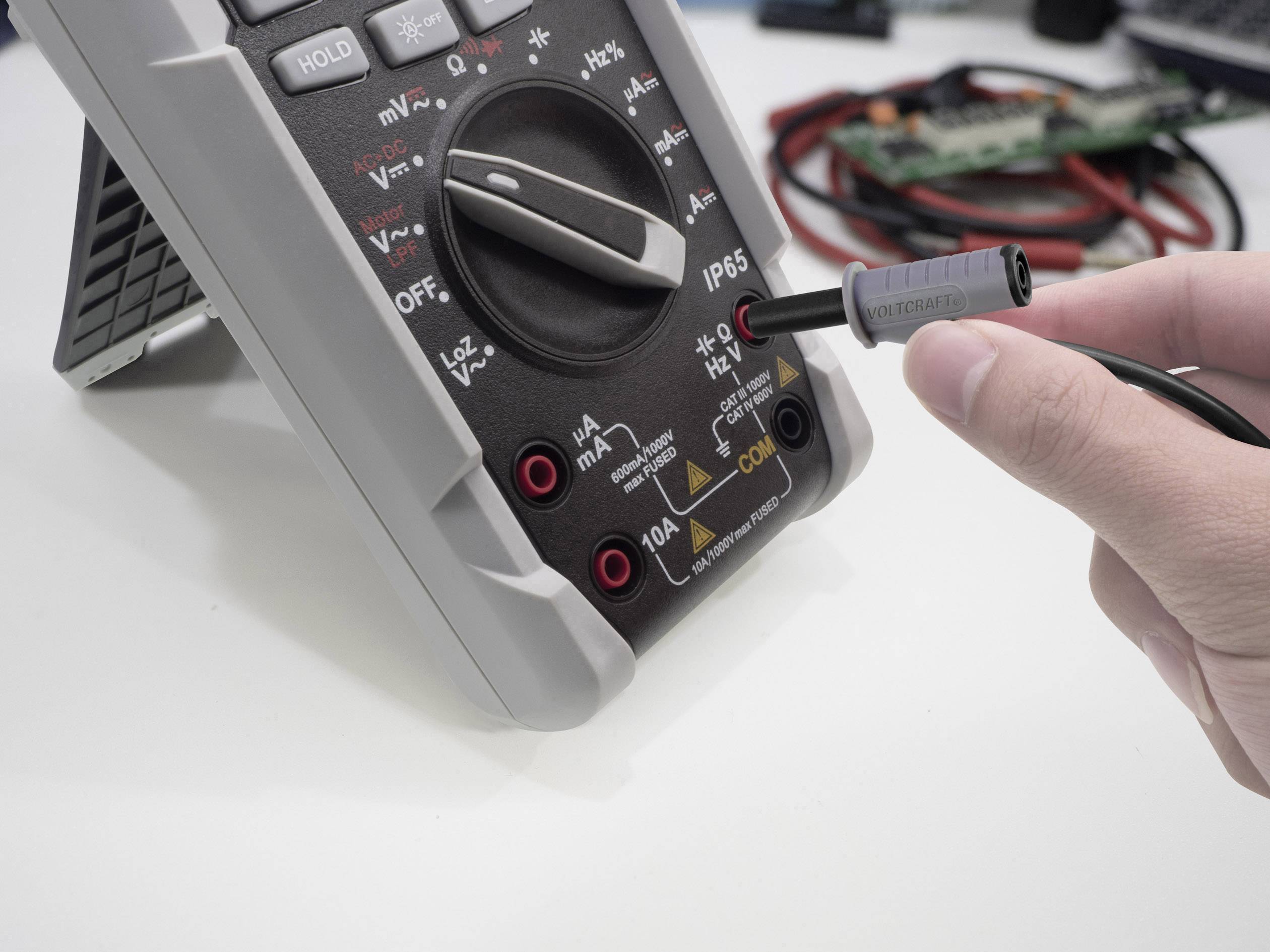 A multimeter set to measure alternating current; one hand is holding a test probe connected to a socket.