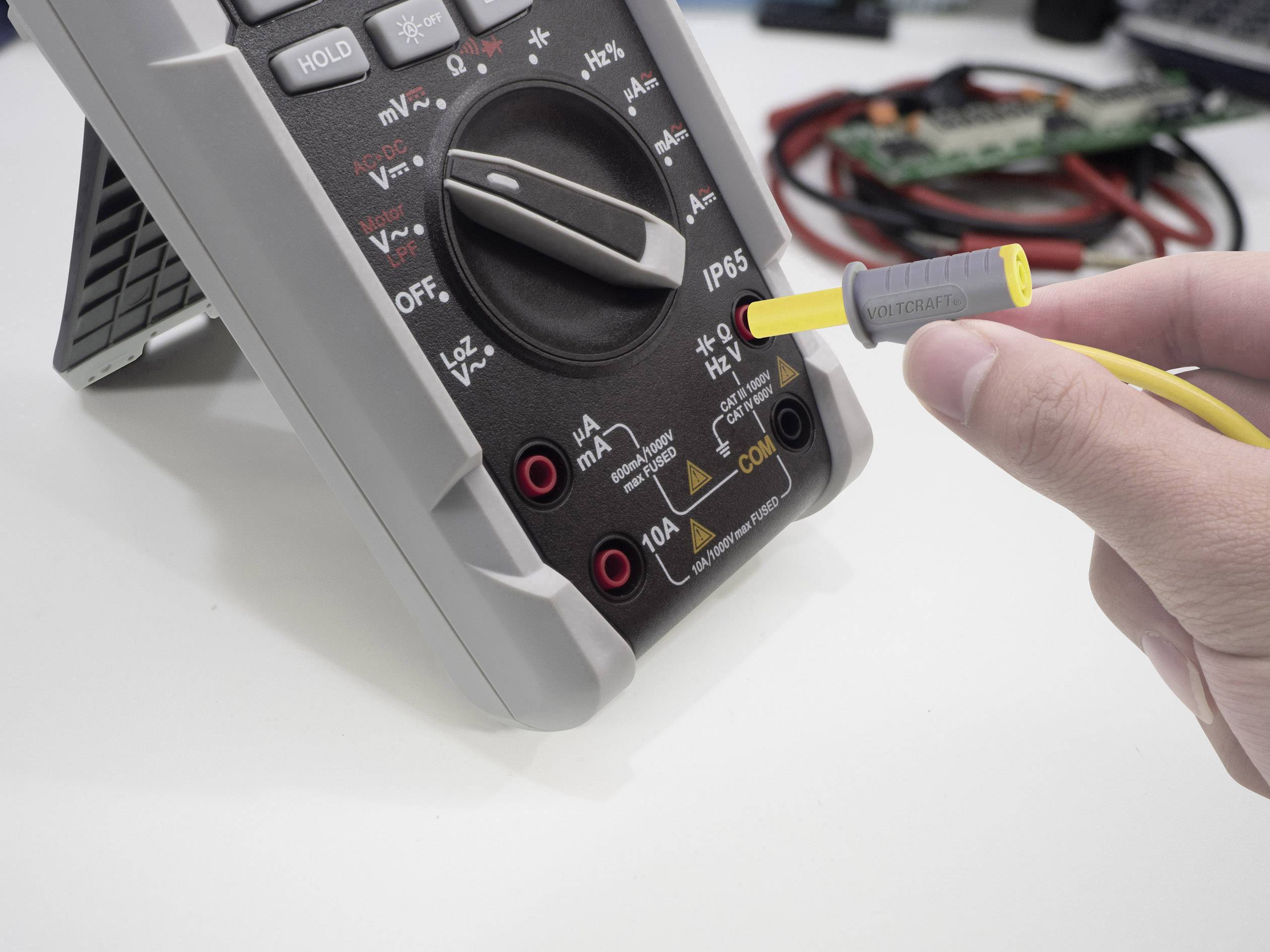 A digital multimeter is being used on a table. One hand is holding a measuring cable that is plugged into the voltage input of the device.