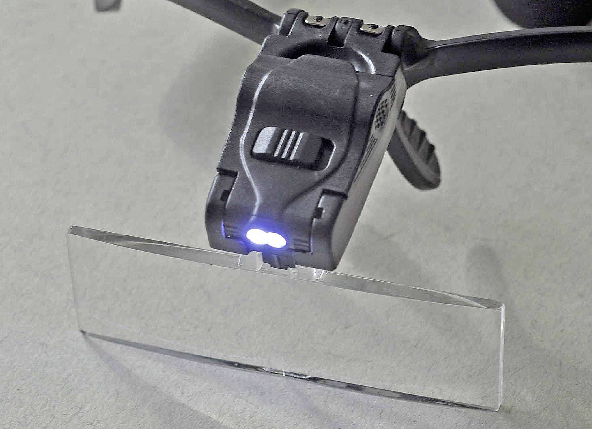 A compact reading light is attached to a pair of spectacle frames. It illuminates a transparent surface underneath.