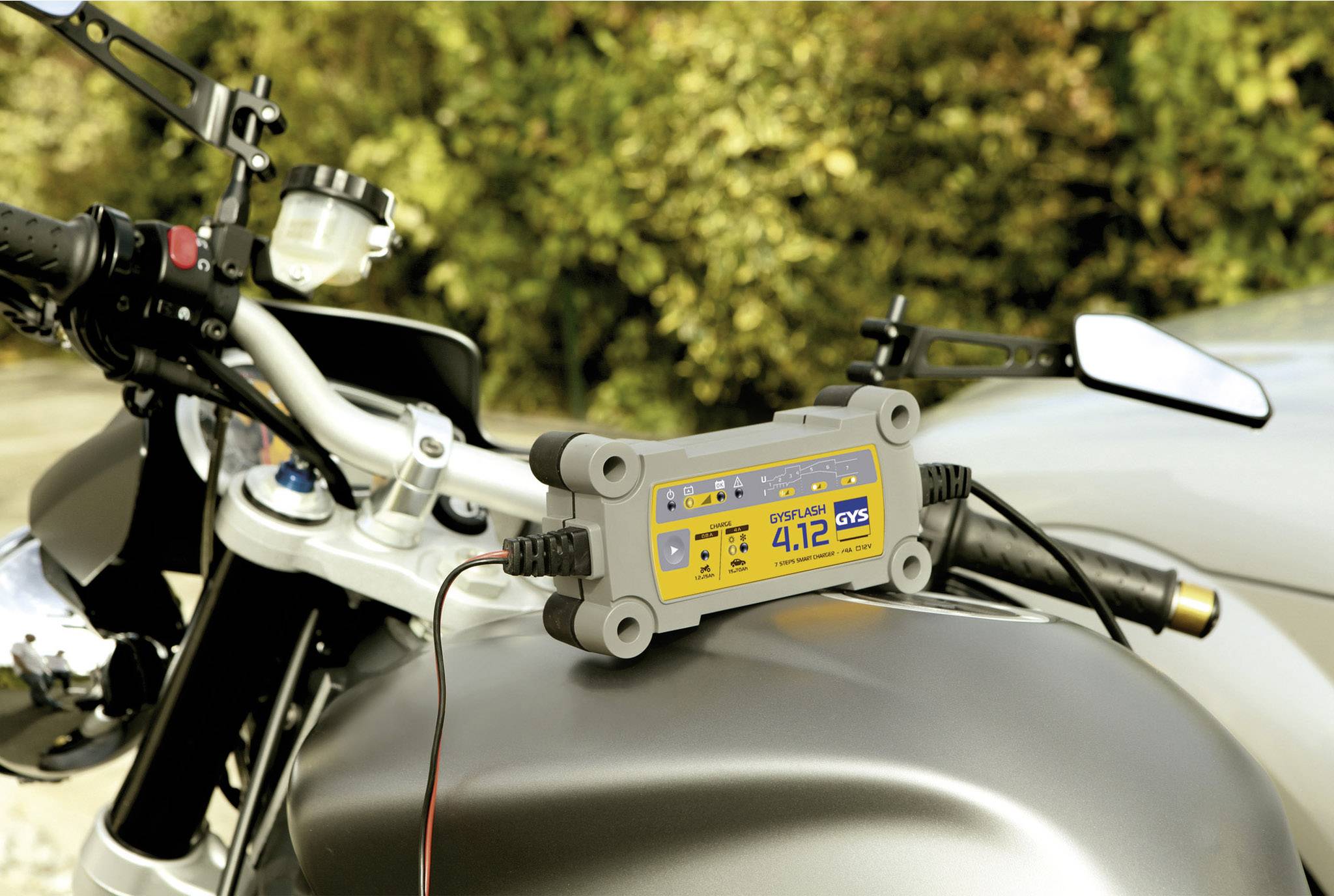 A battery charger is connected to a motorcycle parked on a road with trees in the background.