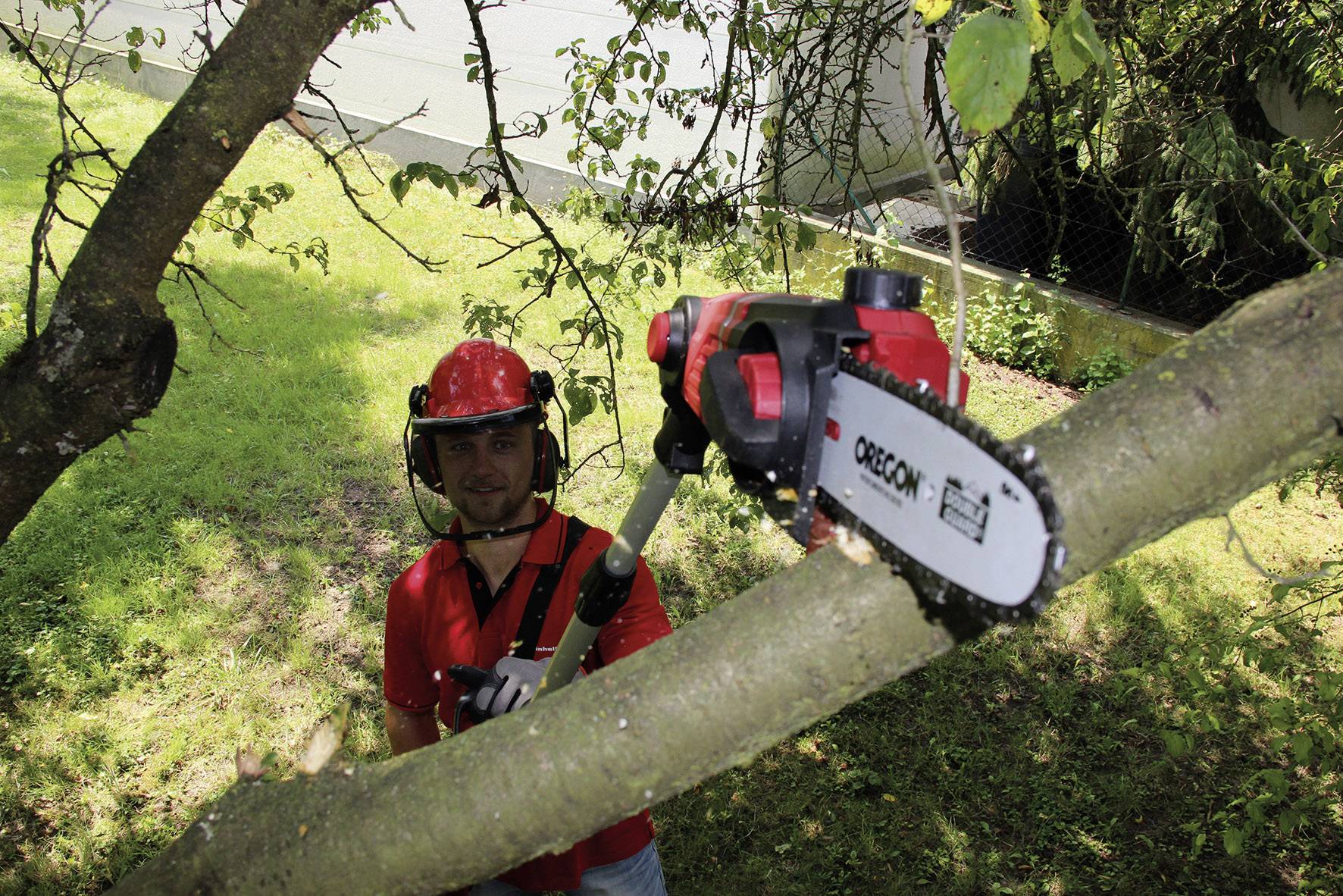 A man wearing a red safety helmet and glasses is cutting a thick branch from a tree using an electric pruning saw.