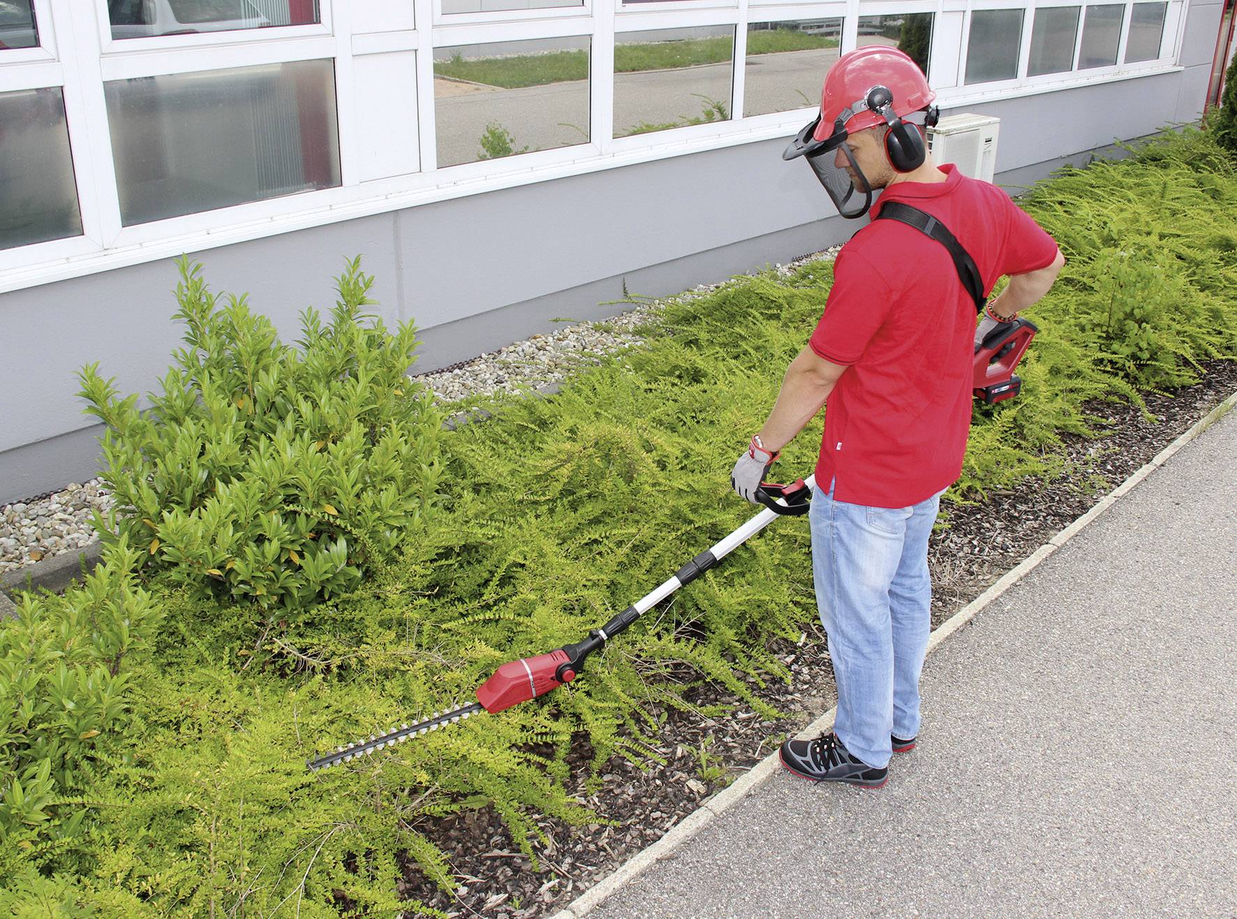 A person in protective clothing is trimming bushes with an electric hedge trimmer in front of a building.