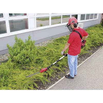 A person in protective clothing is trimming bushes with an electric hedge trimmer in front of a building.