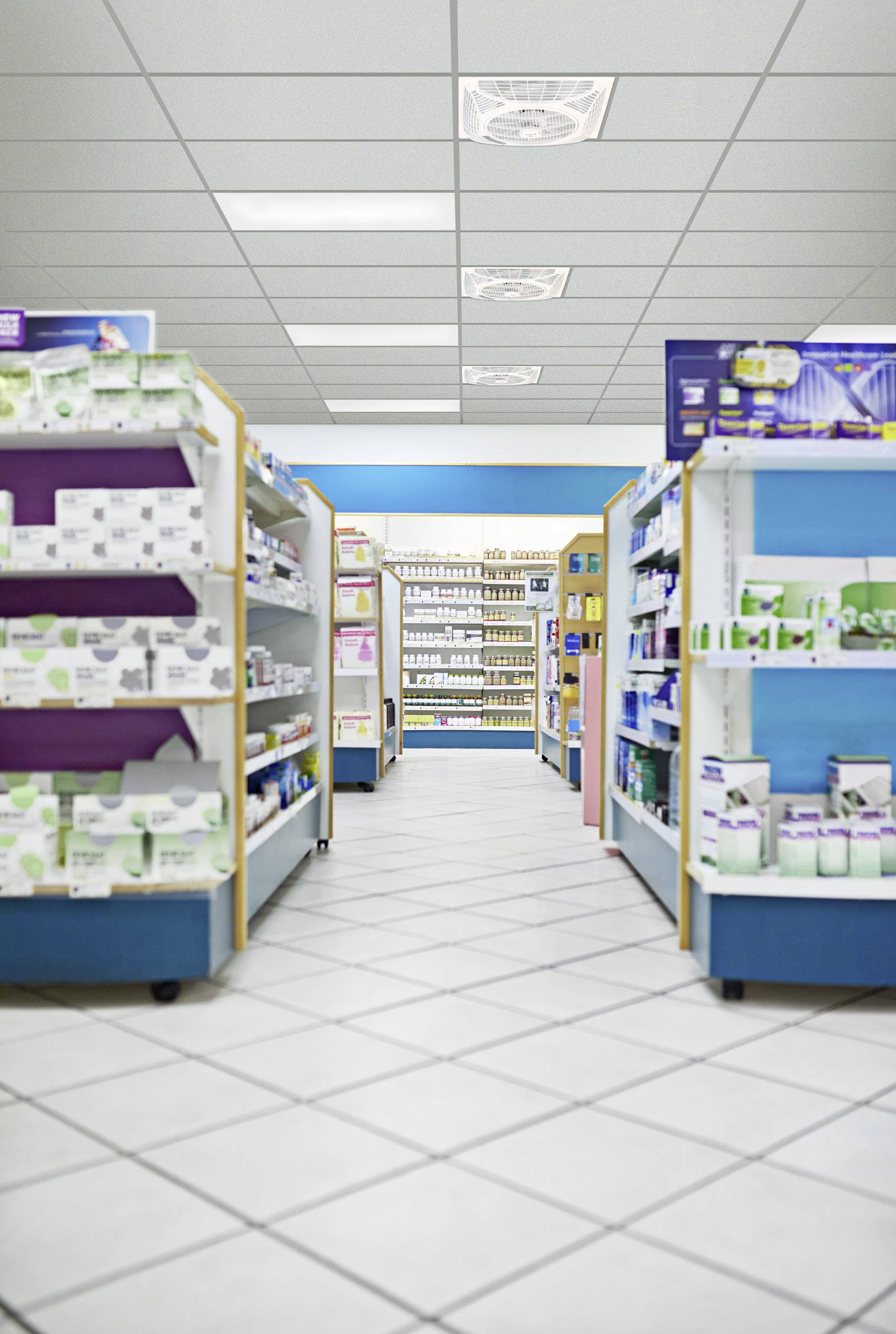 Pharmacy with shelves full of medicines and products stretching back to a counter in the background. Bright lighting and white tiled floors.