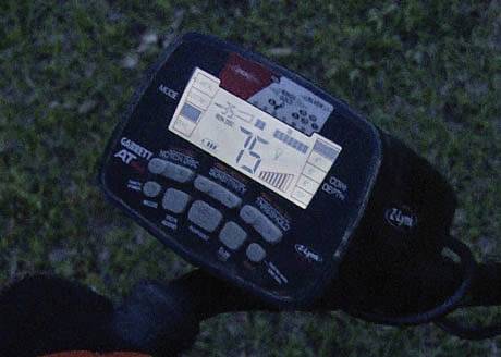 A metal detector displays the number '76' on its screen, indicating the detected metal, with various control buttons underneath.