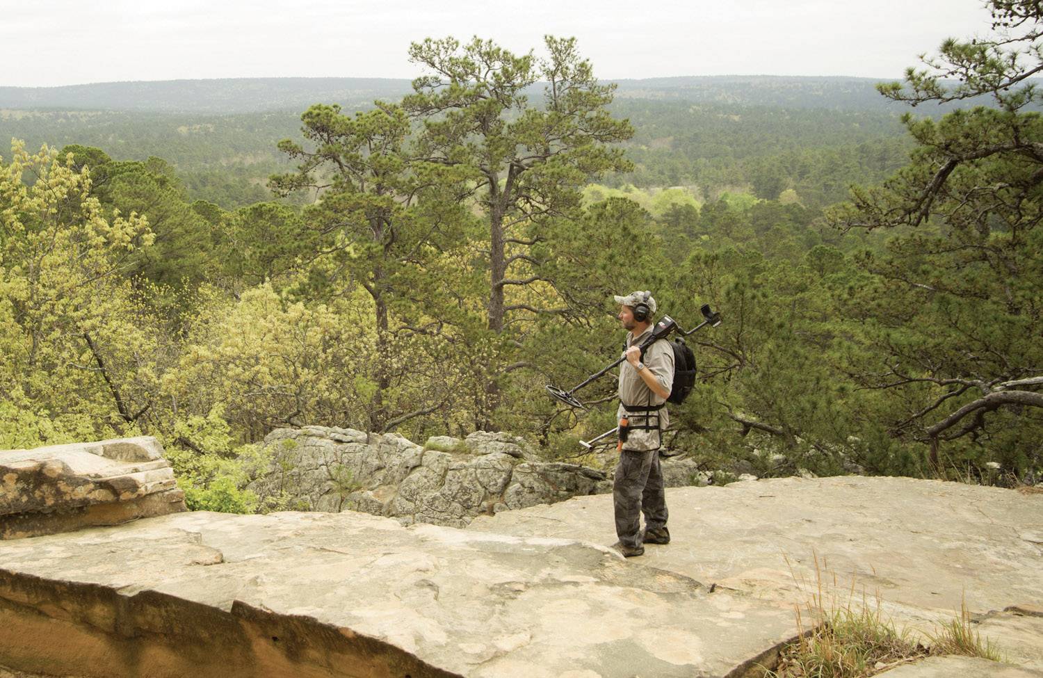A man with a rucksack and camera equipment stands on a rocky outcrop, overlooking a vast, forested national park.