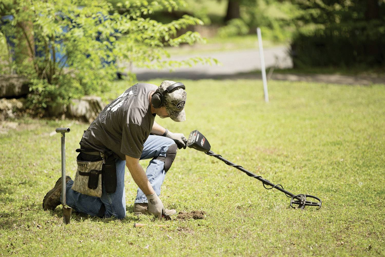 A person is searching for treasures in a meadow using a metal detector. She is kneeling and using tools to explore the ground.
