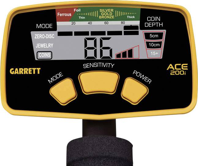 The image shows the front of a metal detector with a digital display. It displays the mode, sensitivity, battery status, and depth.