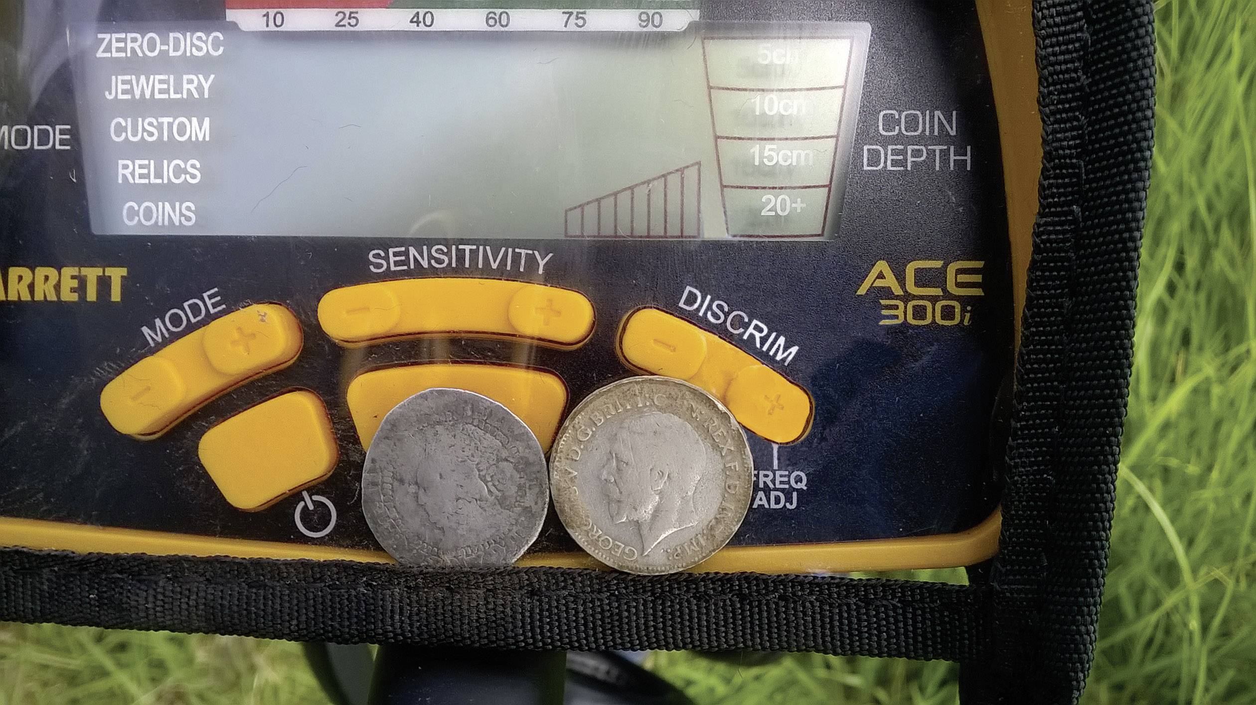 A metal detector display shows reading settings. Beneath it are two old coins. Grass is visible in the background.