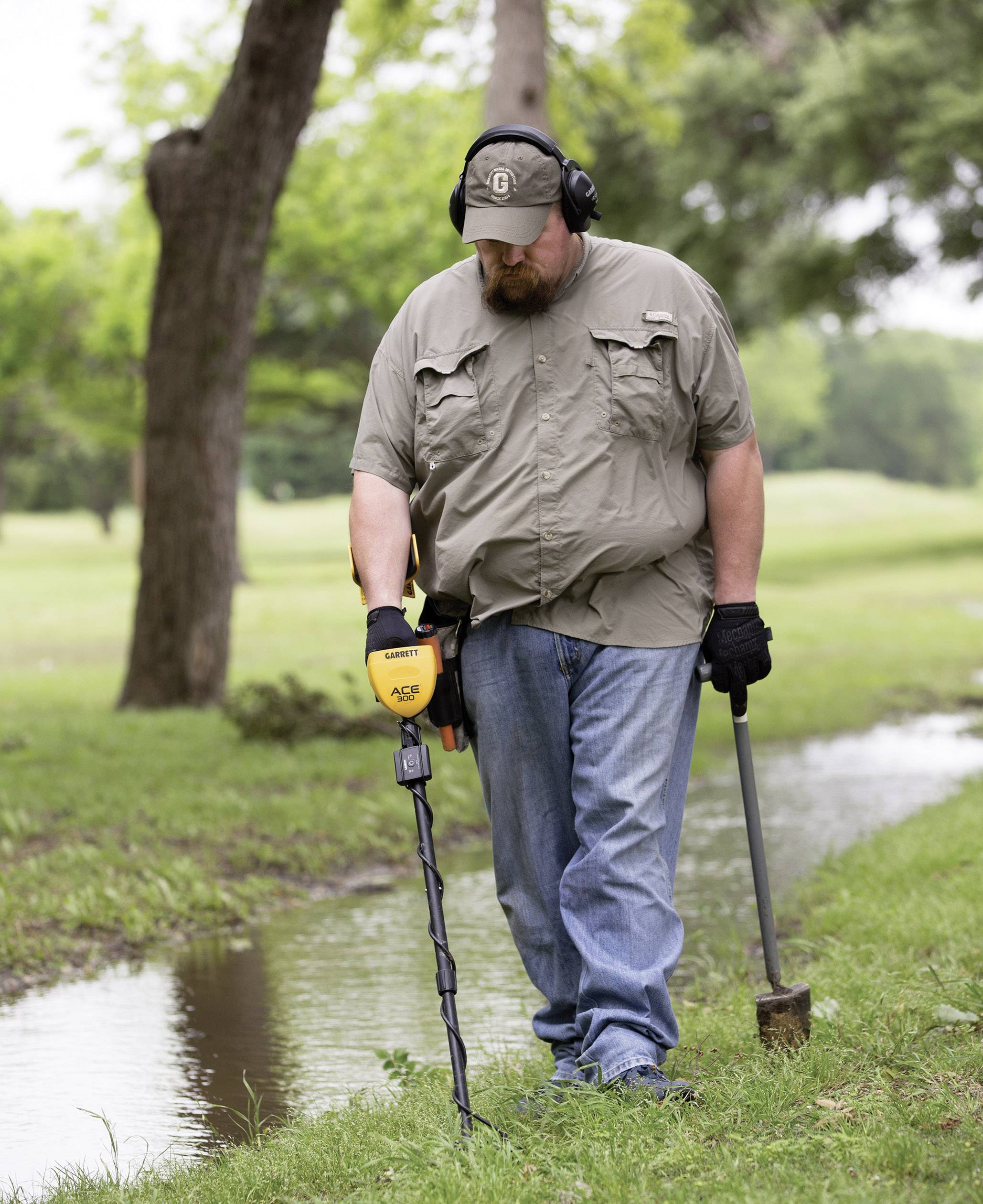 A man is searching for metal with a metal detector beside a small drainage ditch in a green area; he is wearing headphones and gloves.