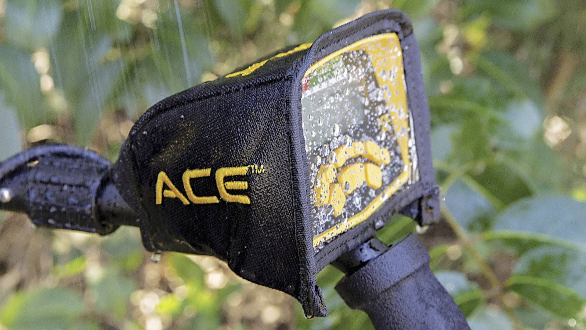 A metal detector with display is drenched in water. Symbols and a scale are visible on the display. Plants in the background.