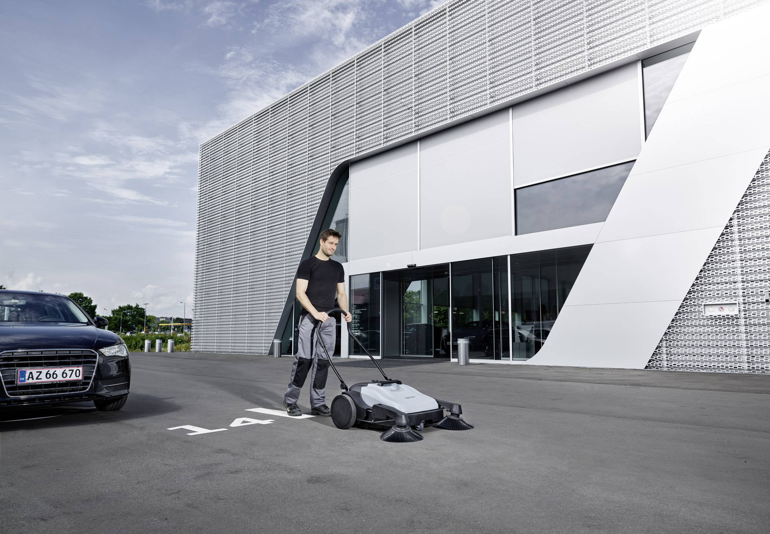 A man is using a mechanical floor cleaner on a car park in front of a modern building on a clear day.